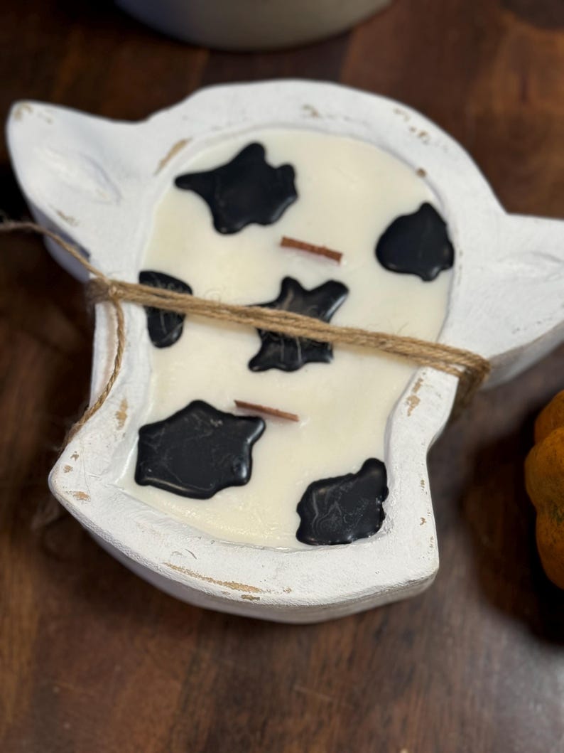 Peut inclure: Un bougeoir blanc en forme de vache avec une bougie blanc cr&egrave;me &agrave; taches noires. La bougie a deux m&egrave;ches en bois et est attach&eacute;e avec de la ficelle. Le support a une finition vieillie.