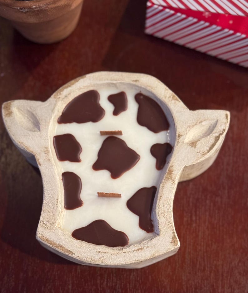 Peut inclure: Une bougie en forme de vache avec une base de cire blanche et des taches marron, plac&eacute;e dans un support en bois blanc cass&eacute; vieilli. La bougie comporte deux m&egrave;ches en bois et est con&ccedil;ue pour ressembler &agrave; une t&ecirc;te de vache. Un objet de d&eacute;coration unique.