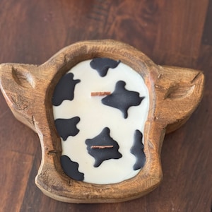 Peut inclure: Une bougie en forme de vache avec un support en bois. La bougie a une surface blanche avec des taches noires de vache et une m&egrave;che en bois. Le support en bois est de couleur marron clair.