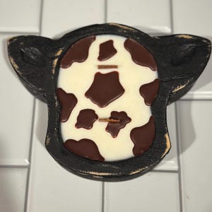 Peut inclure: Un bougeoir noir en forme de vache avec une bougie blanche orn&eacute;e de taches marron. La bougie a deux m&egrave;ches en bois. Le support a une finition vieillie, lui donnant un aspect rustique.