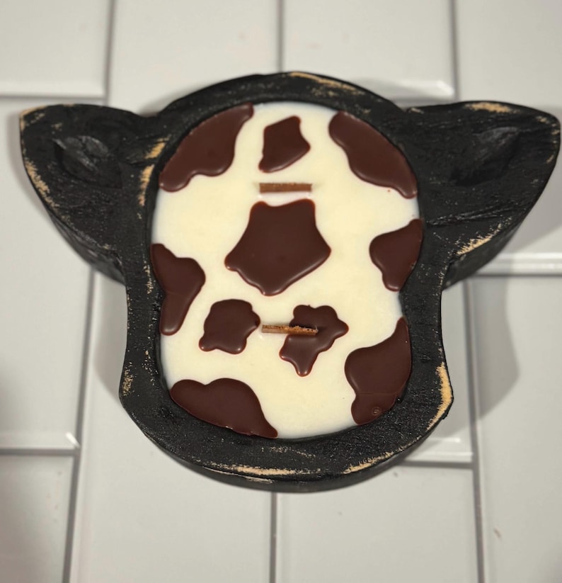 Peut inclure: Un bougeoir noir en forme de vache avec une bougie blanche orn&eacute;e de taches marron. La bougie a deux m&egrave;ches en bois. Le support a une finition vieillie, lui donnant un aspect rustique.