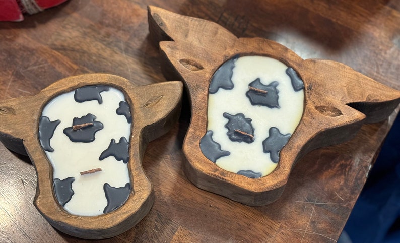 Peut inclure: Deux bougeoirs en bois en forme de vache. Chaque bougeoir contient une bougie blanche cr&eacute;meuse avec des taches noires de vache. Les bougies ont chacune deux m&egrave;ches. Les bougeoirs en bois sont de couleur marron moyen et pr&eacute;sentent des d&eacute;tails sculpt&eacute;s.