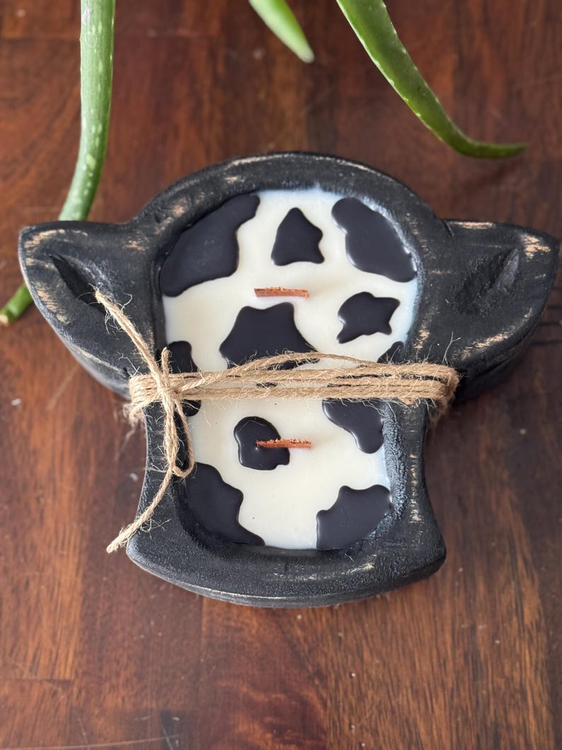 Peut inclure: Un bougeoir en bois noir en forme de t&ecirc;te de vache, rempli de cire blanche et de taches noires. La bougie a deux m&egrave;ches en bois et est attach&eacute;e avec de la ficelle. Le fond est une surface en bois fonc&eacute;.