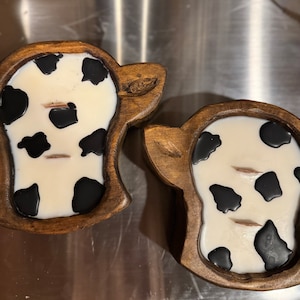 Peut inclure: Deux bougies en forme de vache avec une base blanche et des taches noires sont plac&eacute;es dans des supports en bois. Les supports sont sculpt&eacute;s pour ressembler &agrave; une t&ecirc;te de vache, avec des d&eacute;tails d'oreilles. Les bougies ont deux m&egrave;ches chacune.