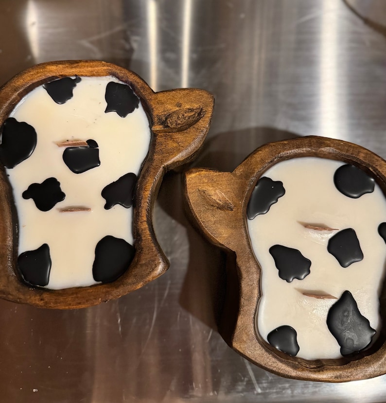 Peut inclure: Deux bougies en forme de vache avec une base blanche et des taches noires sont plac&eacute;es dans des supports en bois. Les supports sont sculpt&eacute;s pour ressembler &agrave; une t&ecirc;te de vache, avec des d&eacute;tails d'oreilles. Les bougies ont deux m&egrave;ches chacune.