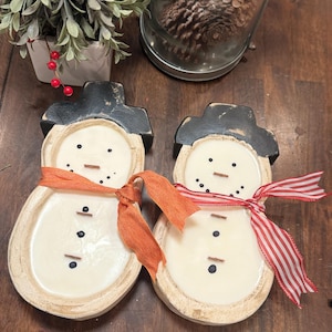 May include: Two snowman-shaped wooden decorations with black top hats and orange and red striped scarves. The snowmen have black dot eyes and button details. The decorations are on a wooden surface.