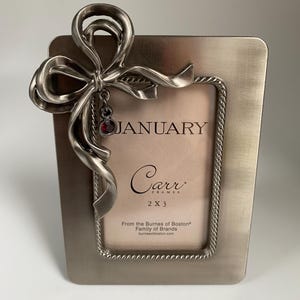 May include: A rectangular silver-toned metal picture frame with a decorative bow and a dangling red gem. The frame has a rope-like border and the word "JANUARY" is visible. The frame is 2 x 3 inches.