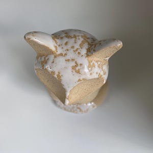 May include: A ceramic fox ornament with a cream glaze and speckled brown detailing. The fox has pointed ears and a rounded head. The figurine is a decorative item, suitable for display on a shelf or table.