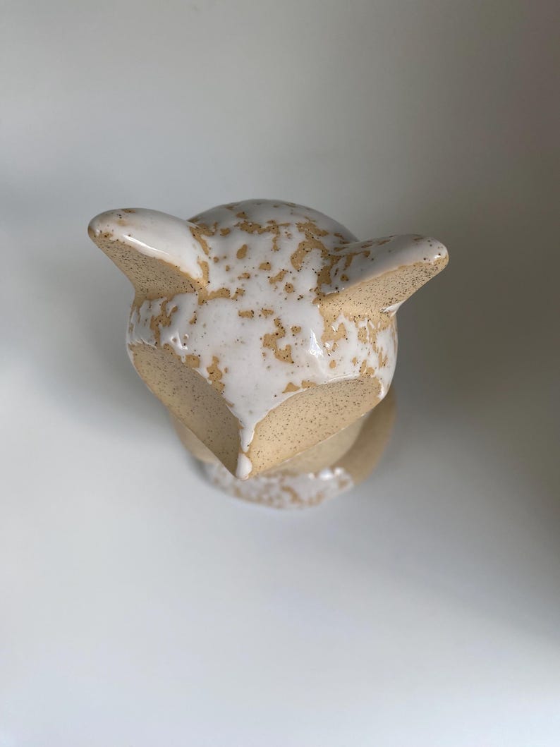 May include: A ceramic fox ornament with a cream glaze and speckled brown detailing. The fox has pointed ears and a rounded head. The figurine is a decorative item, suitable for display on a shelf or table.