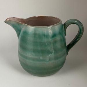 May include: A green ceramic pitcher with a light brown rim and a rounded body. The pitcher has a smooth, glossy finish and a swirling pattern.