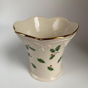 May include: Cream-colored vase with a scalloped rim and gold trim. The vase is decorated with green holly leaves and red berries. A decorative bow is near the top. The vase has a flared shape and a round base.