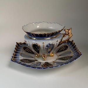 May include: A vintage teacup and saucer set. The cup is white with a blue and gold design, featuring a scalloped rim and a gold handle. The saucer is square with a matching blue and gold pattern. The set is likely porcelain.