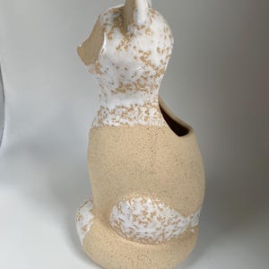 May include: A ceramic fox-shaped vase with a speckled beige and white glaze. The fox is seated, with a hollowed-out back for holding flowers or other decorative items. The ears and tail are detailed with white glaze.