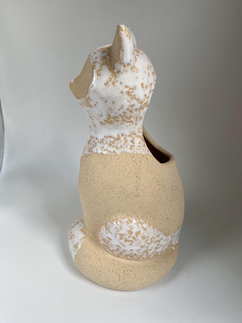 May include: A ceramic fox-shaped vase with a speckled beige and white glaze. The fox is seated, with a hollowed-out back for holding flowers or other decorative items. The ears and tail are detailed with white glaze.