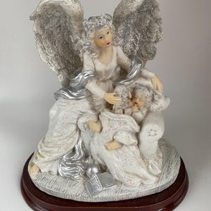 May include: A white angel figurine with silver glitter wings, a white dress, and a halo. The angel is seated and cradling two smaller figures. The base is a dark brown, rounded shape.