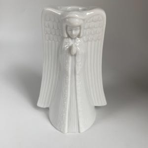 May include: White ceramic angel candle holder. The angel is depicted with folded hands and large wings. The candle holder is designed to hold a single candle. The angel is wearing a long robe with a detailed pattern.