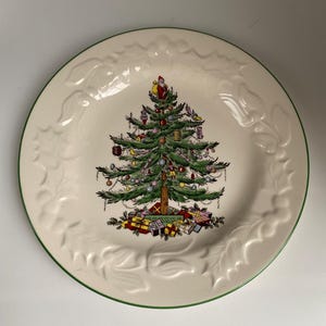 May include: A round, off-white ceramic plate with a green rim and embossed holly leaf design. The plate features a detailed illustration of a decorated Christmas tree with gifts at the base. Santa Claus is at the top of the tree.