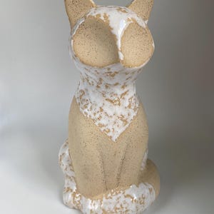 May include: A ceramic cat ornament in a seated pose. The cat has a cream-coloured body with speckled tan accents. The face and chest feature white and tan speckled patterns. The ears are pointed and tan.