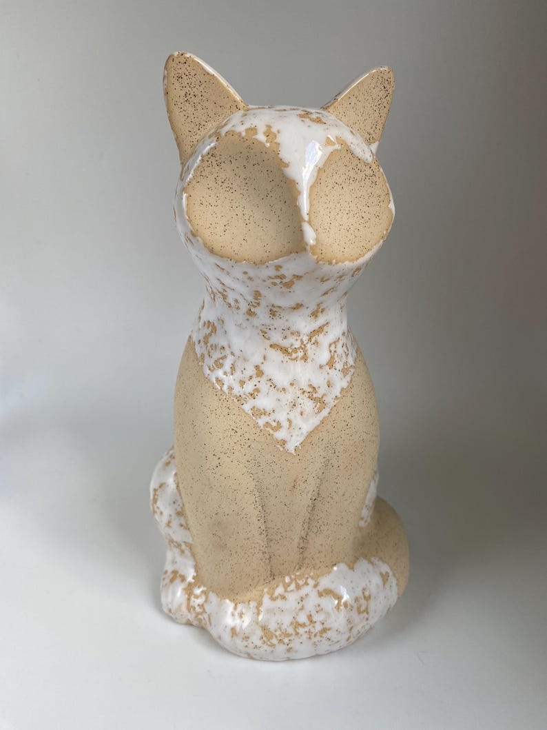 May include: A ceramic cat ornament in a seated pose. The cat has a cream-coloured body with speckled tan accents. The face and chest feature white and tan speckled patterns. The ears are pointed and tan.