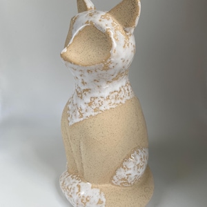 May include: A ceramic fox ornament in a seated pose. The fox features a speckled beige body with white detailing around the face, ears, and tail. The ornament has a smooth, glossy finish.