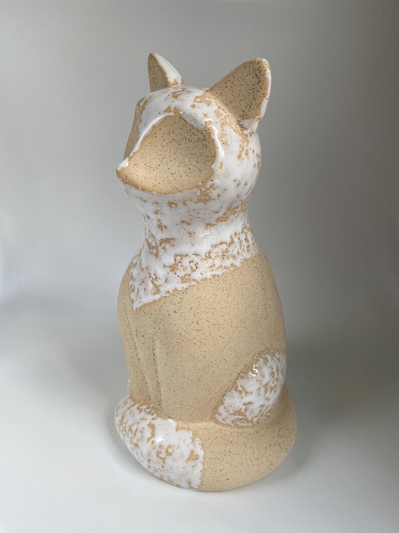 May include: A ceramic fox ornament in a seated pose. The fox features a speckled beige body with white detailing around the face, ears, and tail. The ornament has a smooth, glossy finish.