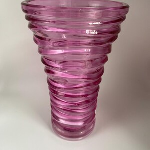 May include: A translucent pink glass vase with a wide, flared rim and a tapered body. The exterior surface features a continuous, spiraling ribbed texture that winds from the narrow base up to the top, creating a series of horizontal ridges.