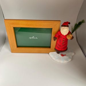 May include: A wooden picture frame with a green interior and the Hallmark logo, next to a Santa Claus figurine holding a small tree branch. The Santa figurine is wearing a red coat and hat, with a white beard. A small red bird is at his feet.