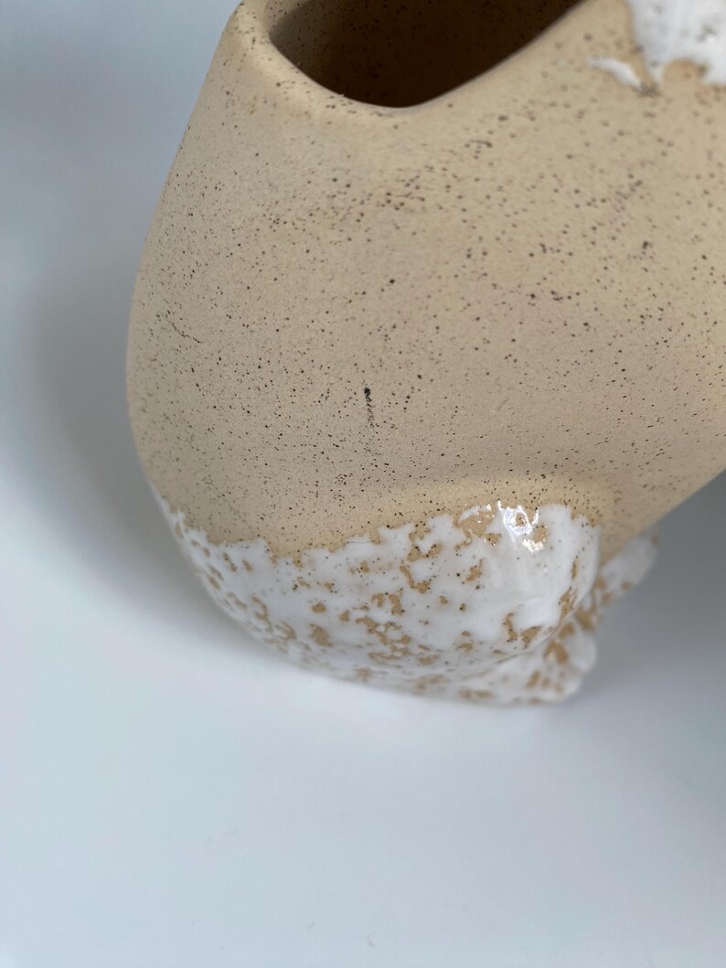 May include: Close-up of a beige ceramic vase with a speckled texture and a white, textured base. The vase has a rounded shape with a smooth, curved opening.