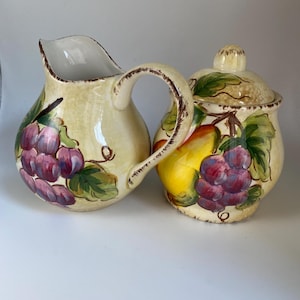 May include: Cream-colored ceramic pitcher and lidded sugar bowl set. Both pieces are decorated with purple grapes, green leaves, and a yellow pear. The pitcher has a curved handle and a spout. The sugar bowl has a rounded shape.