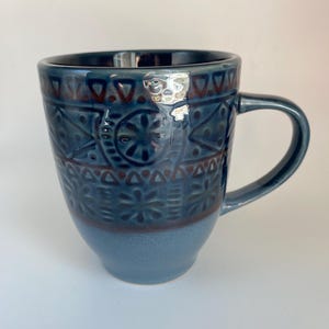 May include: A blue ceramic mug with a handle, featuring a detailed embossed pattern of geometric shapes and floral designs. The mug has a dark blue rim and handle, with a lighter blue base.