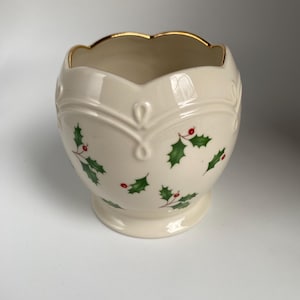 May include: Cream-colored ceramic vase with a scalloped gold rim. The vase features a decorative embossed design and is adorned with green holly leaves and red berries, perfect for holiday decor.