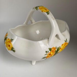 May include: White ceramic basket with a handle and three small feet. The basket is decorated with yellow rose floral patterns. The handle and the sides of the basket feature the floral design. The basket has a scalloped rim.