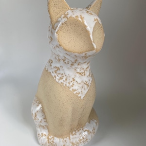 May include: A ceramic cat figurine in a seated position. The cat has a beige body with speckled details and a white glaze that drips down from the head and chest. The ears are pointed, and the tail curls around the body.