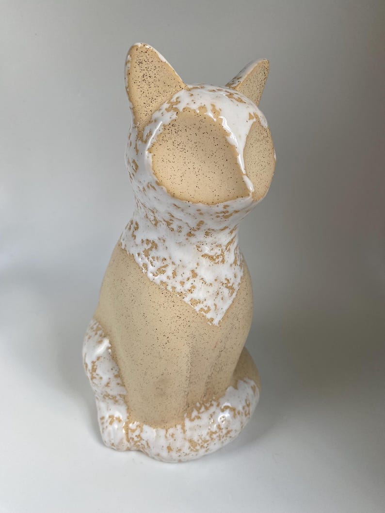 May include: A ceramic cat figurine in a seated position. The cat has a beige body with speckled details and a white glaze that drips down from the head and chest. The ears are pointed, and the tail curls around the body.