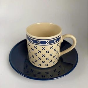 May include: A cream-colored ceramic coffee cup with a blue floral pattern and a matching saucer. The cup has a handle and a blue band around the top. The saucer is a solid dark blue.