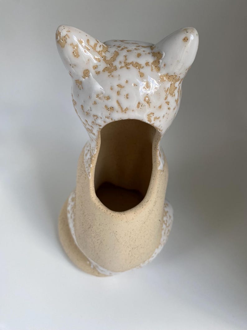 May include: A ceramic fox-shaped planter in a sitting position. The planter is mainly beige with white accents and a speckled, textured finish. The fox's ears and head are white with brown speckles, and the interior is hollow.