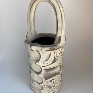 May include: Off-white ceramic vase with a handle. The vase features a floral and leaf design in a darker, contrasting color. The handle is arched and attached to the top of the vase. The interior of the vase is dark.