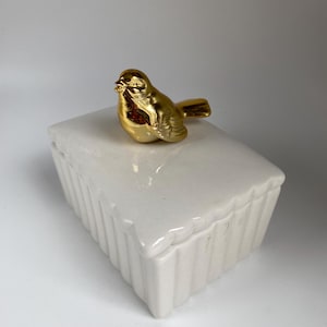 May include: A rectangular white ceramic box with a fluted side design and a removable lid. A small, shiny gold-colored bird figurine is attached to the top of the lid, serving as a handle for the container. The object has a smooth, glossy finish.