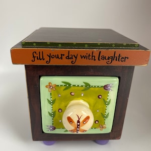 May include: A small, wooden decorative box with a drawer. The drawer front is light green with a butterfly knob. The box is dark brown with purple feet. The top of the box has the text "fill your day with laughter".