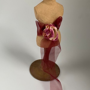 May include: A small, beige fabric mannequin with a burgundy ribbon tied around the waist. The ribbon is adorned with a dried pink and white flower. The mannequin is standing on a gold metal base.