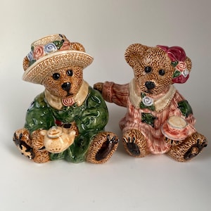 May include: Two ceramic teddy bear figurines. One bear wears a green jacket and a hat with floral accents, holding a small teapot. The other bear is in a pink striped dress and a floral hair accessory, holding a teacup. Both have brown fur.
