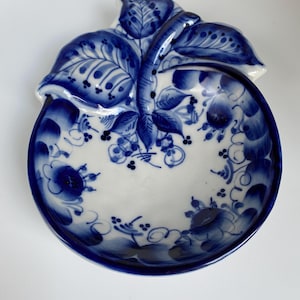 May include: A small, white ceramic dish shaped like an apple with a blue and white floral design. The dish is decorated with a traditional Russian folk art style.