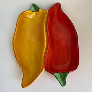 May include: A ceramic serving dish shaped like two connected chili peppers. One side is bright yellow, the other is red. Both have green stems. The dish is likely used for serving appetizers or snacks.