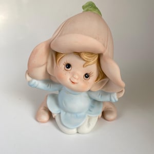 May include: A ceramic figurine of a small child wearing a flower hat. The figurine has a light blue outfit, rosy cheeks, and blonde hair. The flower hat is a soft pink with a green stem.