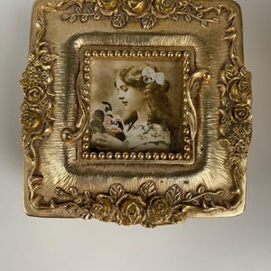 May include: A gold-colored, ornate picture frame with a vintage portrait of a woman holding flowers. The frame is square with a raised floral design and a beaded inner border. The text "size 2" is visible.