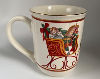 May include: Cream-colored ceramic mug with a gold rim and handle. The mug features a festive design of a child in a red sleigh with a toy horse and Christmas decorations. The design is outlined in red and gold.