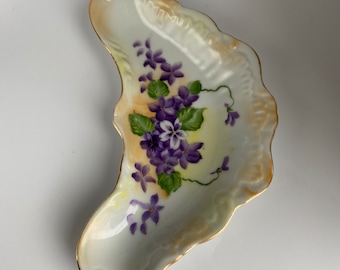 Könnte beinhalten: Eine weiße Porzellanschale mit gewelltem Rand und Goldrand. Die Schale hat die Form eines Blattes und zeigt ein handgemaltes Design aus lila Veilchen mit grünen Blättern.