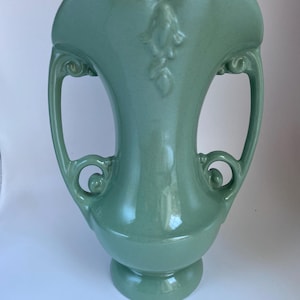 May include: A sage green ceramic vase featuring a flared, heart-shaped rim, two symmetrical scroll-style handles on the sides, and a raised floral relief detail centered on the front. The piece has a smooth, glossy finish and a pedestal base.