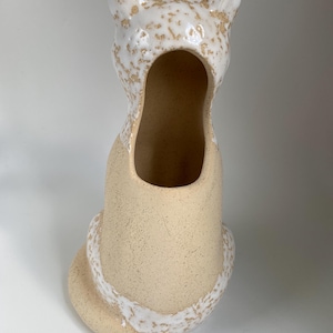 May include: A beige ceramic cat-shaped vase with a textured finish. The head and tail are adorned with a white, speckled glaze. The vase has a large oval opening, and is designed to sit upright.