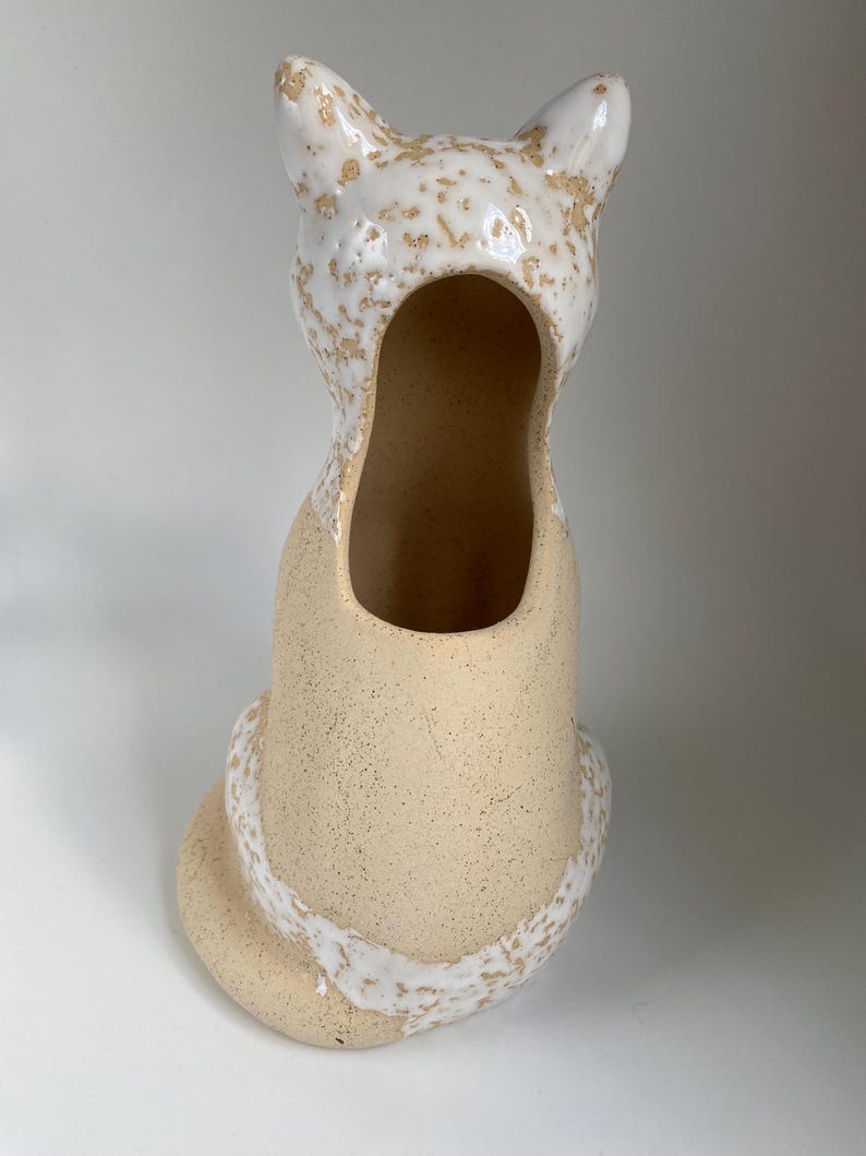 May include: A beige ceramic cat-shaped vase with a textured finish. The head and tail are adorned with a white, speckled glaze. The vase has a large oval opening, and is designed to sit upright.